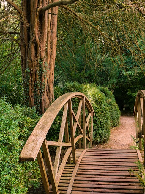 A small wooden footbridge with curved handrails and evenly spaced wooden planks spans over a garden pathway, surrounded by lush green shrubs and dense foliage. In the background, a tall, textured tree